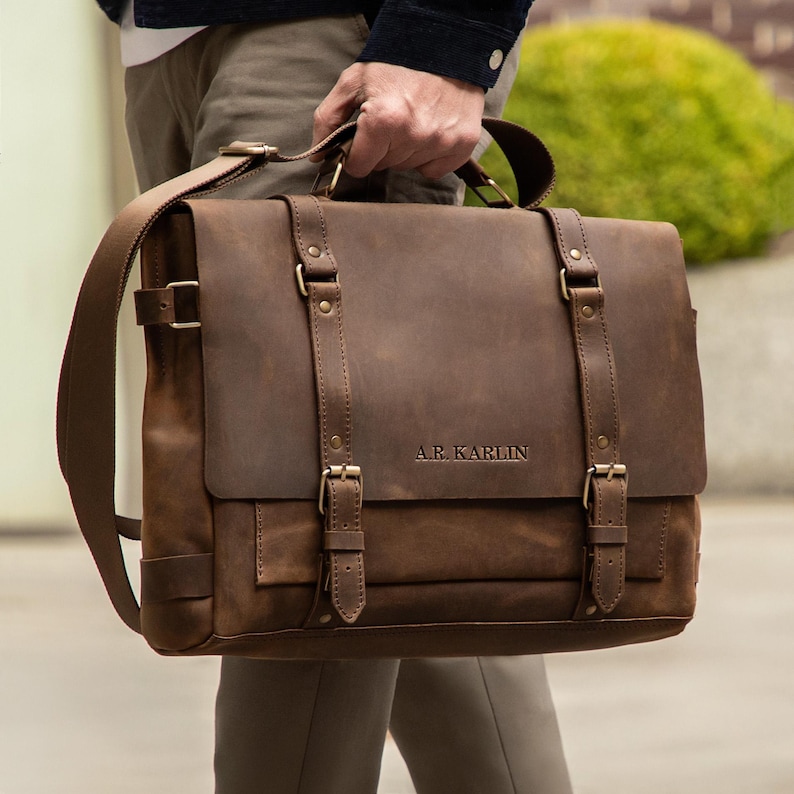 May include: A brown leather satchel with a flap closure, shoulder strap, and top handle. The bag features brass-colored buckles and the text "A.R. Karlin" embossed on the front. The bag is held by a person.