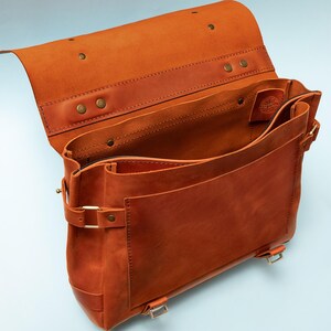 May include: Open, brown leather messenger bag with a flap closure. The bag features multiple compartments, brass hardware, and a buckle closure. The leather has a rich, warm tone, and the bag appears to be well-crafted.