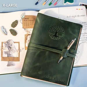 May include: A green leather journal with a tree of life design embossed on the front cover. The journal is closed and has a brown leather strap with a metal buckle. The journal is personalized with the name "ALICE K. REED".