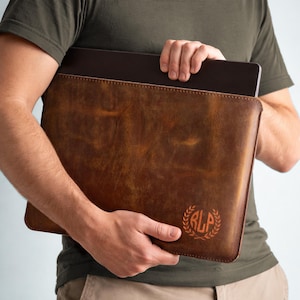 May include: A brown leather laptop sleeve with a monogram design. The sleeve is held by a person wearing a green shirt. The leather has a distressed look and a stitched border. The monogram is in a circular wreath.