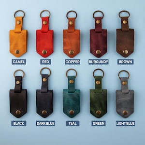 May include: Ten leather keychains in various colors, including camel, red, copper, burgundy, brown, black, dark blue, teal, green, and light blue. Each keychain has a metal ring and a leather strap with a small metal button.