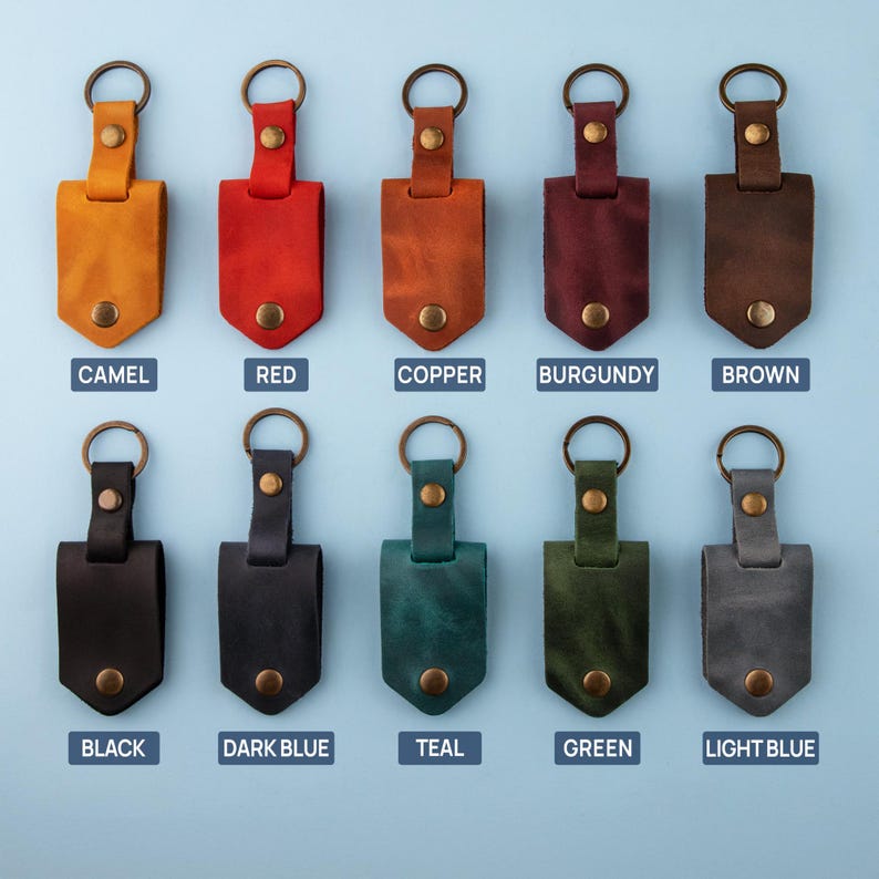 May include: Ten leather keychains in various colors, including camel, red, copper, burgundy, brown, black, dark blue, teal, green, and light blue. Each keychain has a metal ring and a leather strap with a small metal button.