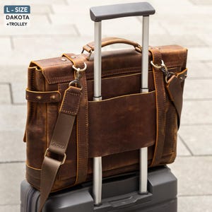 May include: A brown leather briefcase with a shoulder strap and handle, resting on a rolling suitcase. The briefcase has a distressed finish and metal hardware. The text "L-SIZE DAKOTA + TROLLEY" is in the upper left corner.