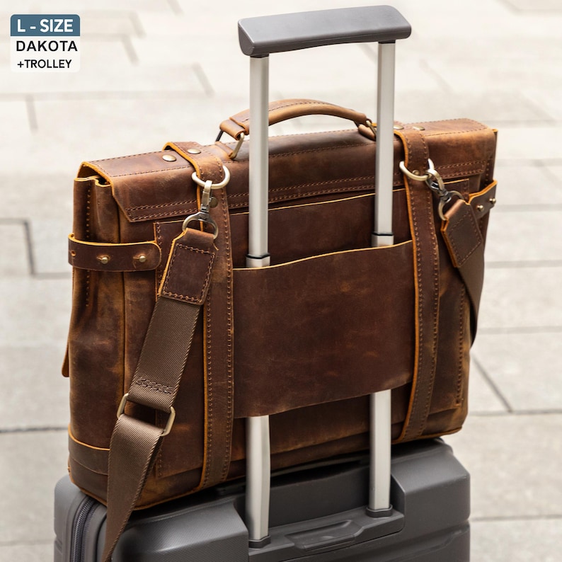 May include: A brown leather briefcase with a shoulder strap and handle, resting on a rolling suitcase. The briefcase has a distressed finish and metal hardware. The text "L-SIZE DAKOTA + TROLLEY" is in the upper left corner.
