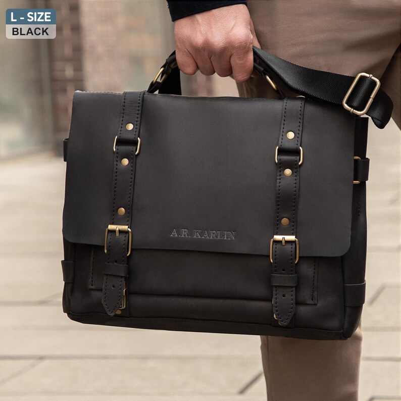 May include: Black leather messenger bag with a flap closure, adjustable shoulder strap, and gold-colored metal hardware. The bag has two front straps with buckles and the text "A.R. Karlin" embossed on the front.