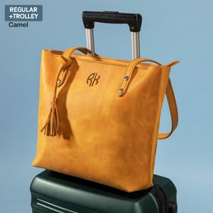 Może przedstawiać: Torebka typu tote ze sk&oacute;ry w kolorze camel z wyhaftowanym monogramem "RK" z przodu. Torebka ma długi pasek i kr&oacute;tszy uchwyt. Torebka leży na zielonej walizce.