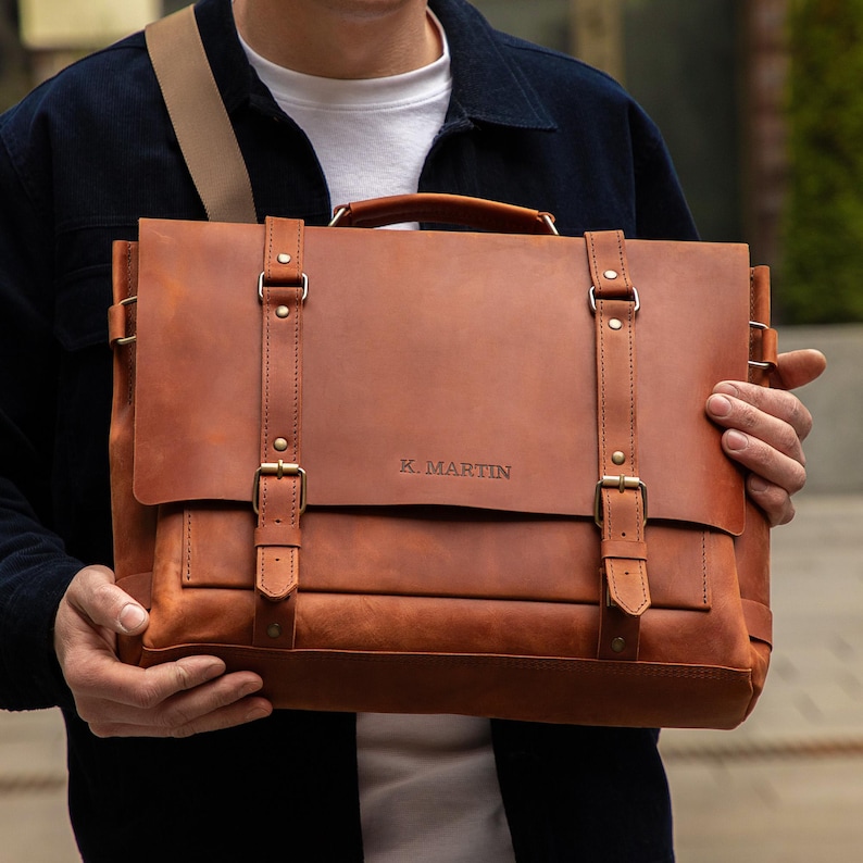 May include: A brown leather briefcase with a top handle and shoulder strap. The bag has two front straps with buckles and the text "K. MARTIN" is visible on the front. The bag is held by a person.