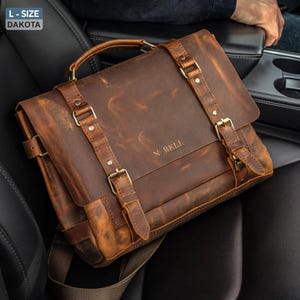 May include: A brown leather messenger bag with a top handle, shoulder strap, and buckle closures. The bag has a distressed finish and the text "N. BELL" is visible on the front. The bag is sitting on a black car seat.