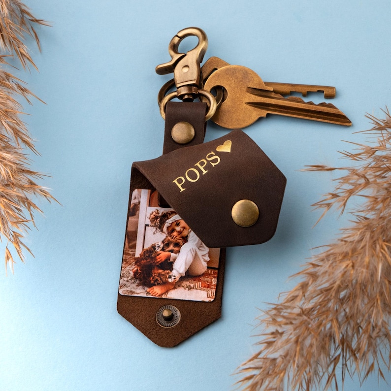 May include: Brown leather photo keychain with a gold metal clasp and a small photo insert. The photo shows a young girl holding a dog. The keychain is engraved with the word "POPS" and a small heart.
