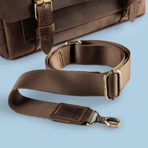 May include: A brown leather satchel with a matching brown nylon shoulder strap. The strap has brass-colored metal hardware, including a swivel clasp and rectangular buckles. The bag and strap are set against a light blue background.