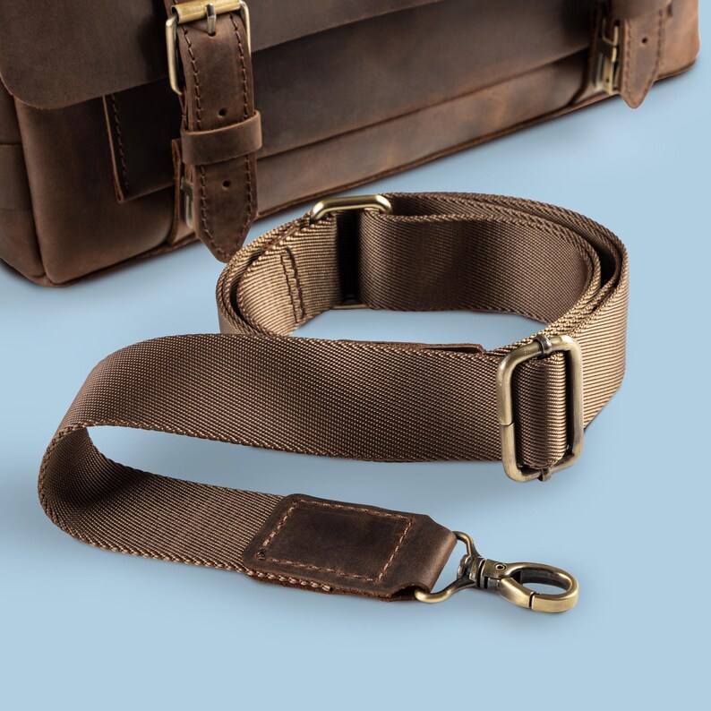 May include: A brown leather satchel with a matching brown nylon shoulder strap. The strap has brass-colored metal hardware, including a swivel clasp and rectangular buckles. The bag and strap are set against a light blue background.