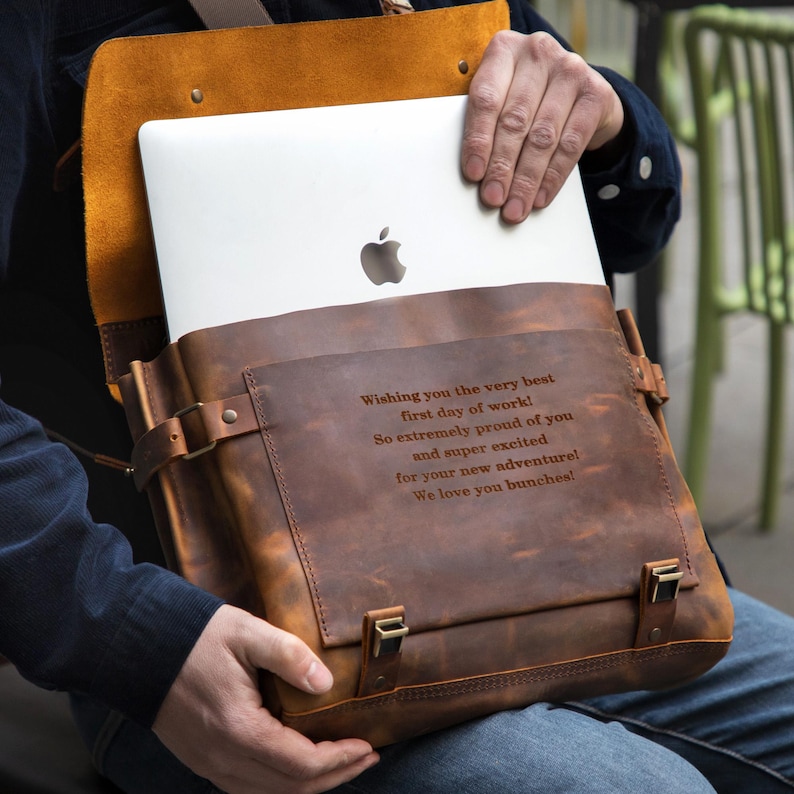 May include: A brown leather messenger bag with a flap closure, holding a laptop. The bag has a distressed finish and metal hardware. Text on the bag reads: "Wishing you the very best first day of work!" and more.
