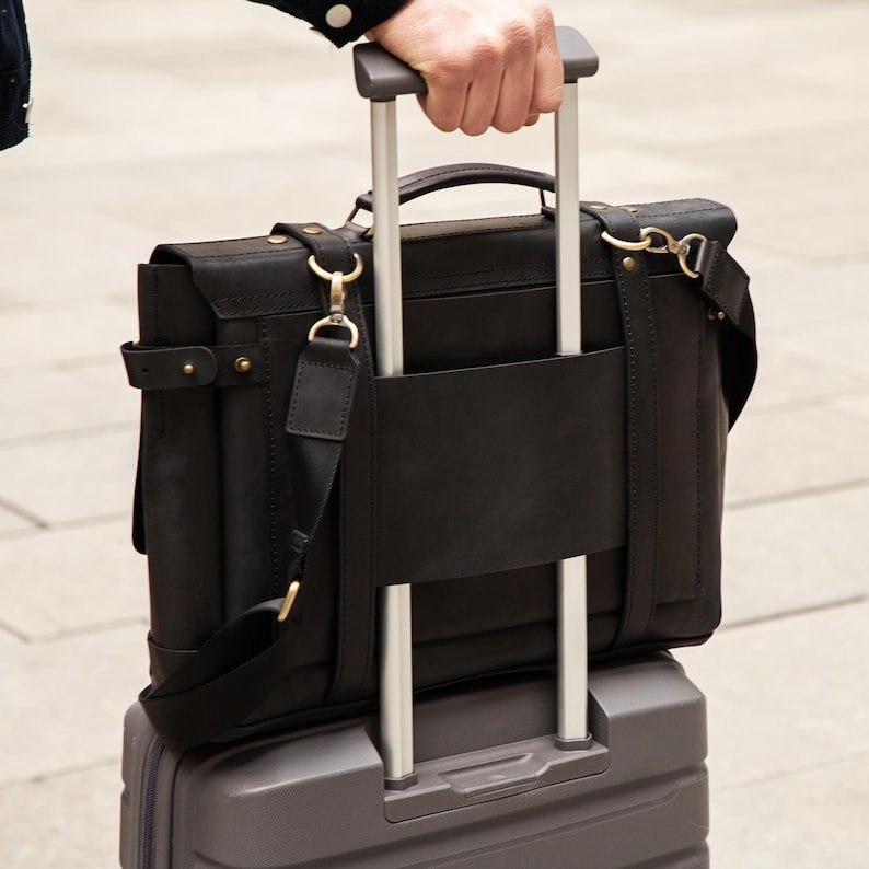 May include: A black leather briefcase with gold-colored hardware is secured to a gray rolling suitcase. The briefcase has a shoulder strap and a top handle. The suitcase has a retractable handle. The image is taken outdoors.