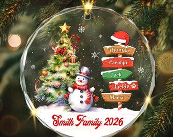 May include: A round, clear glass Christmas ornament featuring a decorated pine tree, a snowman with a red scarf, and a wooden signpost with the names Thomas, Carolyn, Jack, Jackie, and Maria. The text Smith Family 2026 is printed at the bottom.