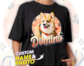 May include: Black t-shirt with a custom photo of a corgi dog wearing a red bandana. The dog's name, "Domino", is written in a retro font on the shirt.