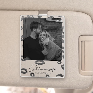 May include: A rectangular car visor accessory with a black and white photo of a couple. The photo is surrounded by a border of lipstick kiss marks. The text "Get home safe I LOVE YOU" is written in cursive below the photo.
