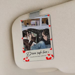 May include: Clear acrylic car visor clip featuring a photo of a couple. The photo is surrounded by red heart accents. The text "Drive safe love. I need you here with me" is written in cursive.