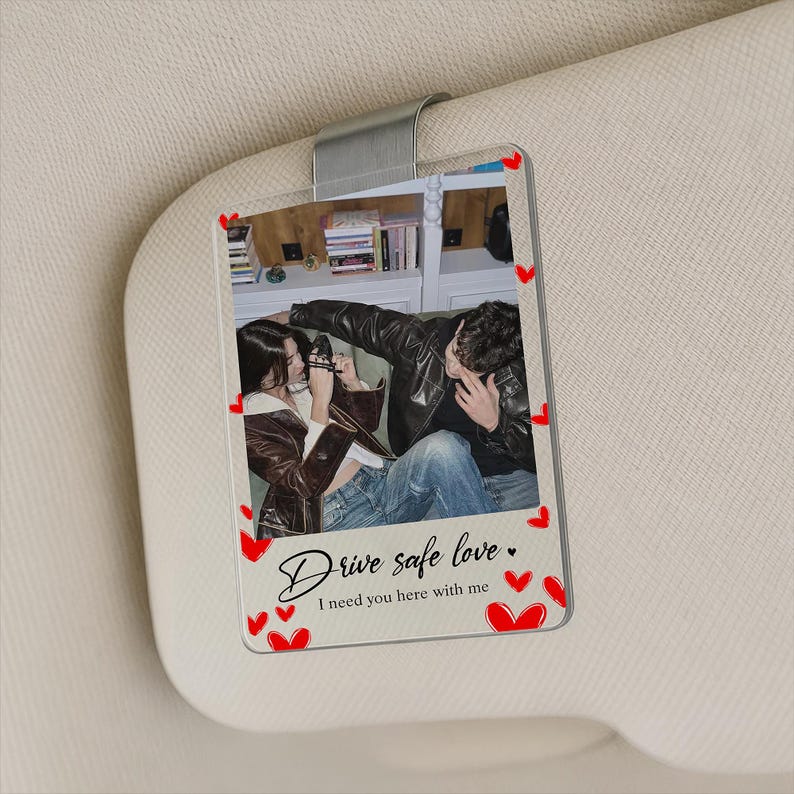May include: Clear acrylic car visor clip featuring a photo of a couple. The photo is surrounded by red heart accents. The text "Drive safe love. I need you here with me" is written in cursive.