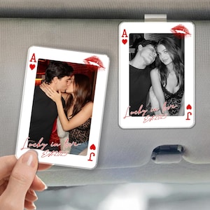 May include: Two playing card-shaped car visor clips. One shows a couple kissing, the other a couple embracing. Both cards have a red lip print and the text "Lucky in love Est.2025". The cards are framed with red hearts and the letter "A" and "J".