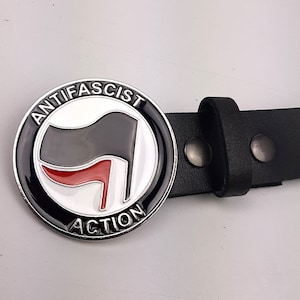Pode incluir: Um cinto de couro preto com uma fivela prateada que apresenta uma bandeira preta e vermelha com as palavras "Anti-Fascist Action" ao redor da borda.