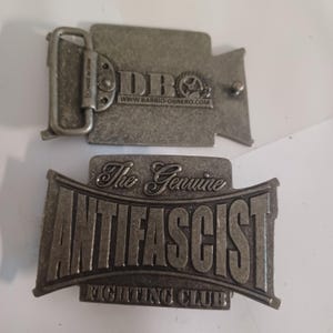 Pode incluir: Uma fivela de cinto de metal prateado com o texto "The Genuine Anti-Fascist Fighting Club" e uma fivela de cinto de metal prateado menor com o texto "DBO www.barrio-obrero.com".