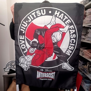 Puede incluir: Bandera negra con un gráfico de Jiu-Jitsu de dos personas en una llave, con las palabras "LOVE JIU-JITSU" y "HATE FASCISM" en un círculo. El texto "The Genuine Antifascist Fighting Club" está debajo.