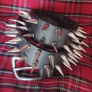 Op de afbeelding: Een zwarte leren riem met zilveren metalen spikes. De riem is dubbelgevouwen en de gesp is zichtbaar.