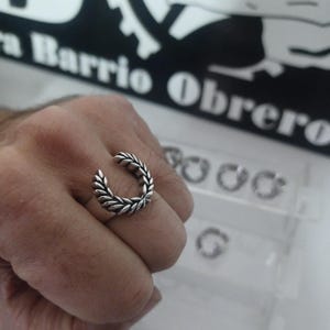 May include: A silver ring with a laurel wreath design. The ring is on a person's finger.