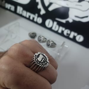 Può includere: Un anello d'argento con un design dettagliato con un emblema centrale circondato da una corona d'alloro. L'anello è su una mano, con altri anelli visibili sullo sfondo. Il testo "Mora Barrio Obrero" è visibile.