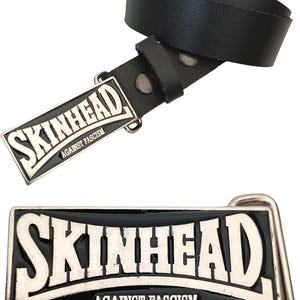 Op de afbeelding: Zwarte leren riem met een zilveren metalen gesp. De gesp heeft de woorden "Skinhead" en "Against Fascism" in een vet, wit lettertype.