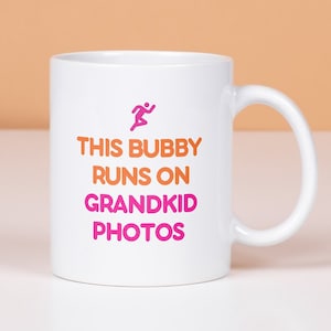 May include: White ceramic coffee mug with a pink and orange design. The mug has a running person icon and the text "This Bubby Runs on Grandkid Photos".