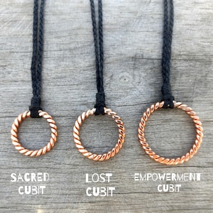 Könnte beinhalten: Drei kupferfarbene Halsketten mit gedrehten Ringanhängern, die an schwarzen Kordeln hängen. Die Halsketten sind mit "Sacred Cubit", "Lost Cubit" und "Empowerment Cubit" beschriftet. Der Hintergrund ist eine verwitterte Holzoberfläche.