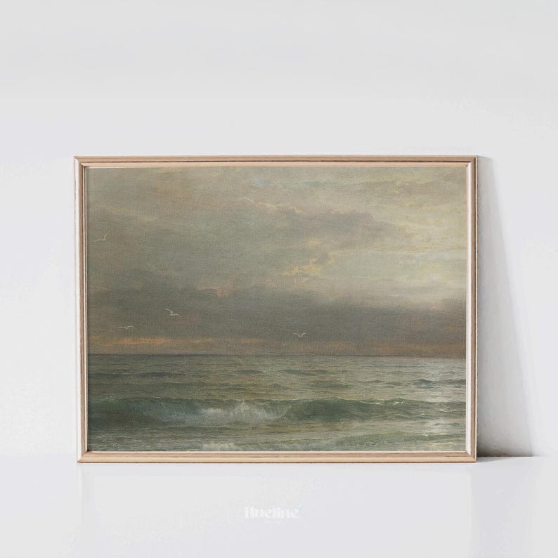 May include: A framed vintage seascape painting. The artwork depicts a moody ocean scene with waves, seagulls, and a cloudy sky. The color palette is muted, with shades of gray, green, and beige. The frame is a light gold color.