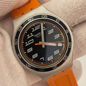 以下が含まれることがあります： 黒の文字盤とオレンジのアクセントが付いたシルバーのSwatch腕時計。ベゼルは黒で白い数字が書かれており、ストラップは黒でオレンジのアクセントが付いている。