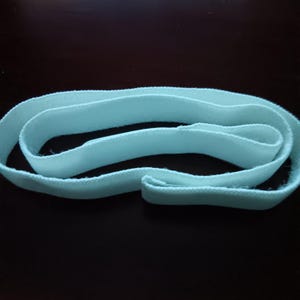 May include: A long, continuous strip of light blue elastic fabric band. The material features a woven, textured surface with visible fibers along the edges. The band is coiled into an oval shape, displaying its flexible and stretchable construction.