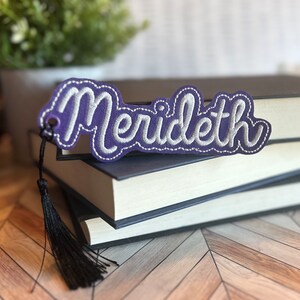 Puede incluir: Marcapáginas de cuero morado con el nombre "Meredith" bordado en hilo blanco. El marcapáginas tiene un borla negra.