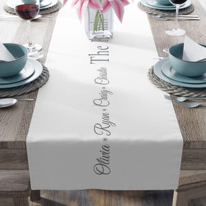 May include: A white table runner with the names "Olivia, Ryan, Craig, Christie, The H" printed vertically in a script font. The runner is set on a wooden table, with place settings and a vase of pink flowers.