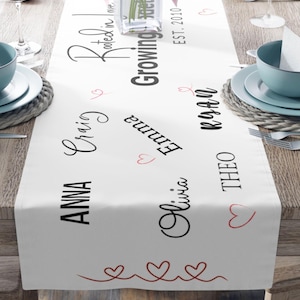 May include: White table runner with black text that reads "Rooted in Love, Growing Together, EST. 2010" and names "Anna, Craig, Emma, Olivia, Theo, Kian". The runner has a red heart design.