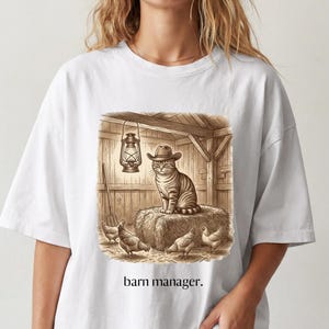 May include: A white t-shirt featuring a sepia-toned graphic of a cat wearing a cowboy hat sitting on a hay bale in a barn with chickens and a lantern. The text "barn manager." is printed in black below the illustration on the front of the shirt.