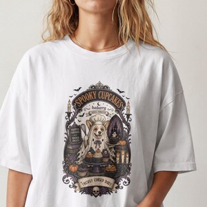 May include: A white t-shirt featuring a graphic print of a dog dressed as a ghost, surrounded by cupcakes, candles, and bats. The text reads "SPOOKY CUPCAKES", "bakery", "HAUNTED CUPCAKES AND WICKED SWEETS", and "FRESHLY CURSED DAILY" with a skull at the bottom.
