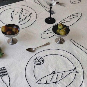 Op de afbeelding: Een witte tafelkleed met zwarte geborduurde illustraties van dining elementen. Het ontwerp omvat borden met eten, bestek en een baguette. Twee kleine metalen kommen bevatten fruit, en een wijnglas staat op de tafel.