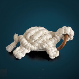 Peut inclure: Un jouet en forme de tortue crocheté en blanc avec un anneau en bois pour la queue. La tortue est couchée sur le ventre, les pattes tendues.