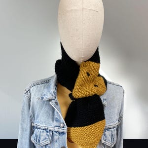 Peut inclure: Écharpe tricotée noire et dorée sur un mannequin. L'écharpe est faite de fil épais, avec des sections noires et dorées alternées. L'écharpe est enroulée autour du cou du mannequin et portée avec une veste en jean.