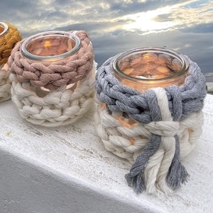 Peut inclure: Trois photophores en verre avec des housses en crochet. Les housses sont de différentes couleurs : rose, gris et blanc. Les bougies sont allumées et brillent.