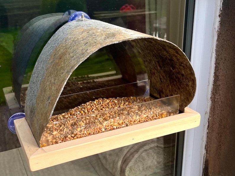 Peut inclure: Mangeoire &agrave; oiseaux en bois avec un toit incurv&eacute; en m&eacute;tal vieilli. La mangeoire a une fa&ccedil;ade en plastique transparent et est remplie de graines pour oiseaux. Elle est fix&eacute;e &agrave; une fen&ecirc;tre avec des ventouses bleues.