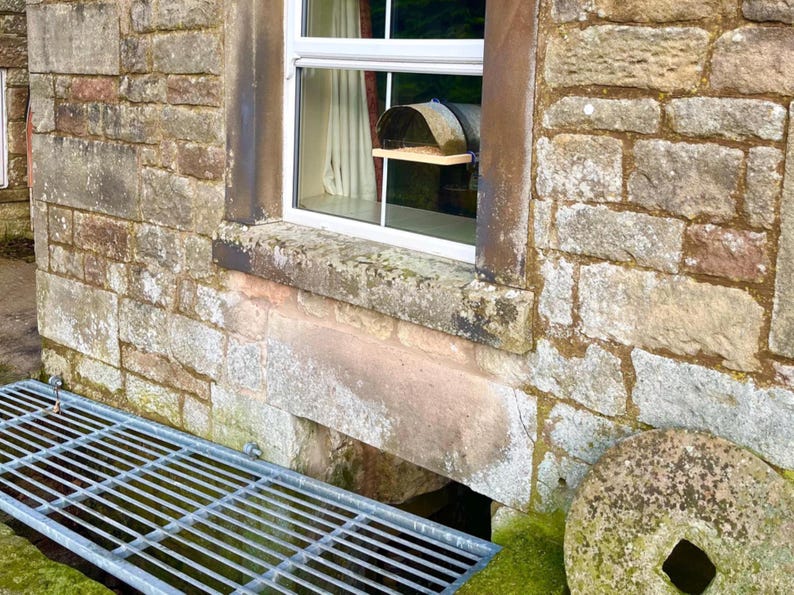 Peut inclure: Vue ext&eacute;rieure d'un b&acirc;timent en pierre avec une fen&ecirc;tre. La fen&ecirc;tre a un cadre blanc et une petite &eacute;tag&egrave;re. Une grille m&eacute;tallique est au premier plan et une meule en pierre est &agrave; droite. Le mur en pierre du b&acirc;timent est patin&eacute;.