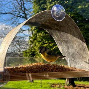 Peut inclure: Un mangeoire &agrave; oiseaux fix&eacute; &agrave; une fen&ecirc;tre, avec un petit oiseau jaune et bleu perch&eacute; &agrave; l'int&eacute;rieur. La mangeoire a une base en bois et un plateau d'alimentation en plastique transparent rempli de graines. Un toit incurv&eacute; en bois vieilli offre un abri.