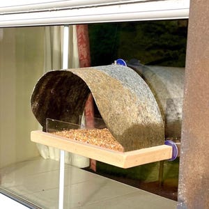 Peut inclure: Un nichoir rustique avec un toit incurv&eacute; et textur&eacute; dans des tons de marron, de gris et de beige. La mangeoire a un plateau en bois rempli de graines pour oiseaux et est mont&eacute;e sur un support blanc. Le design est visible &agrave; travers une fen&ecirc;tre.