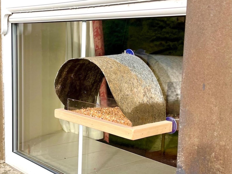 Peut inclure: Un nichoir rustique avec un toit incurv&eacute; et textur&eacute; dans des tons de marron, de gris et de beige. La mangeoire a un plateau en bois rempli de graines pour oiseaux et est mont&eacute;e sur un support blanc. Le design est visible &agrave; travers une fen&ecirc;tre.