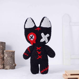 May include: A handmade black crochet doll with white ears and facial details. The doll has red stitched accents and a red and white eye design. The doll is standing upright on a white surface.